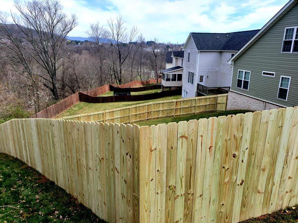 Wooden fence enclosing a yard on a sloped grassy hill, with houses and trees in the background.