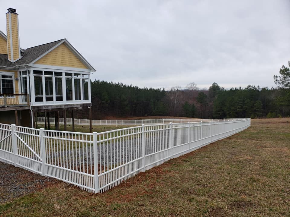 White fence surrounding a yellow house with a porch and windows, bordering a field and woods under a cloudy sky.