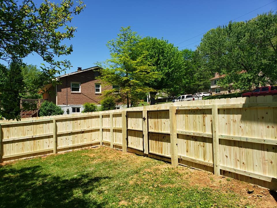 Wooden fence encloses a grassy yard, with a two-door gate. A brick house and trees are in the background under a blue sky.