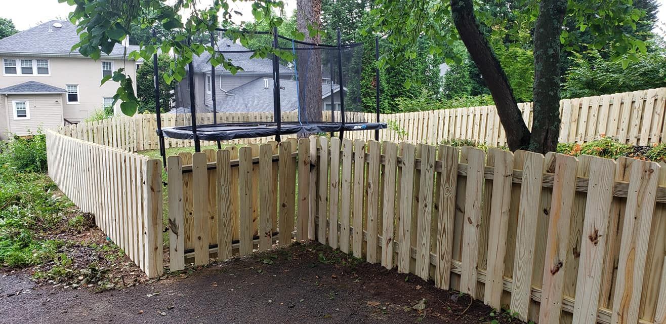 A wooden fence surrounds a yard with a trampoline and a house in the background. Green trees add to the scene.