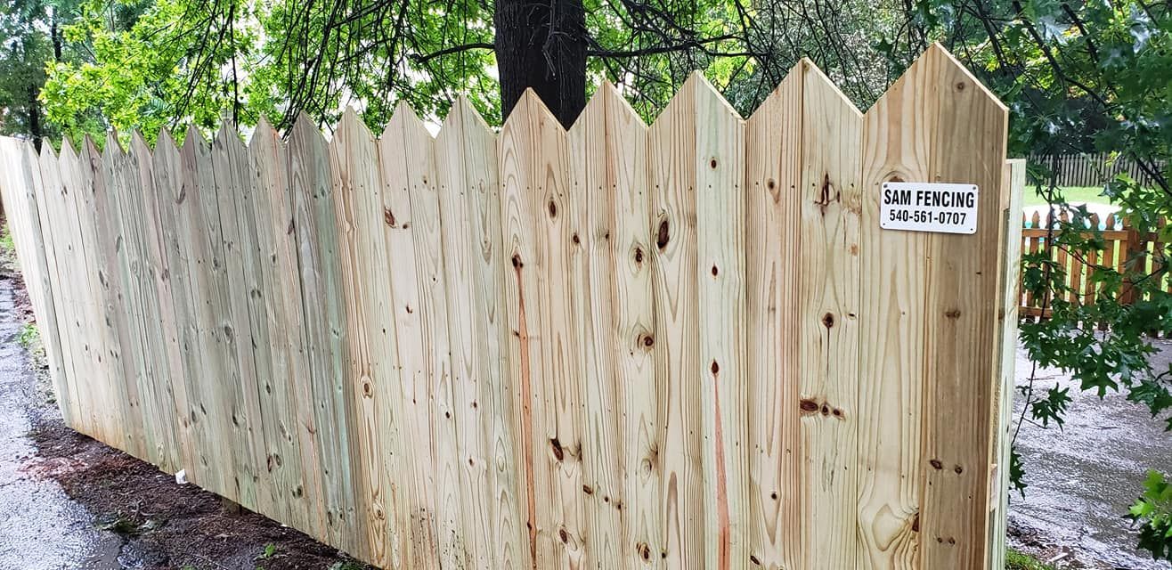A wooden fence with pointed tops stands next to a road, surrounded by trees.