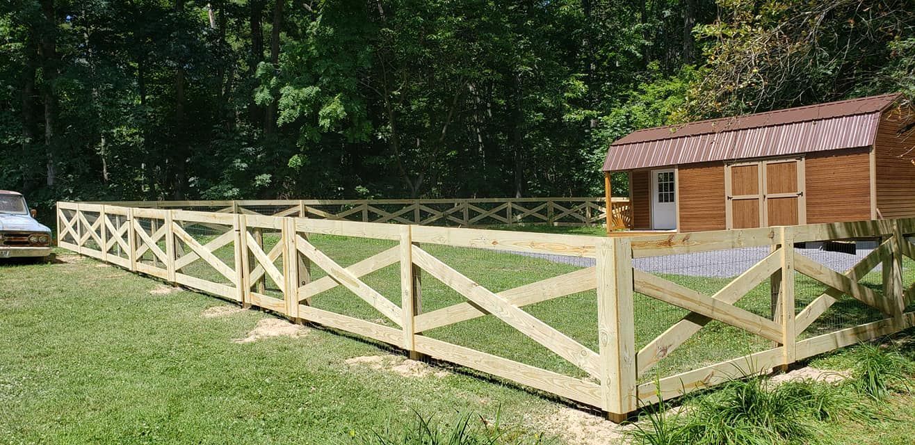Wooden fence surrounds a grass area with a brown shed in the background; green trees and grass.