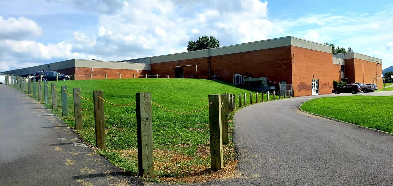 A long, brick building with a gray roof sits behind a grassy hill and a paved road.