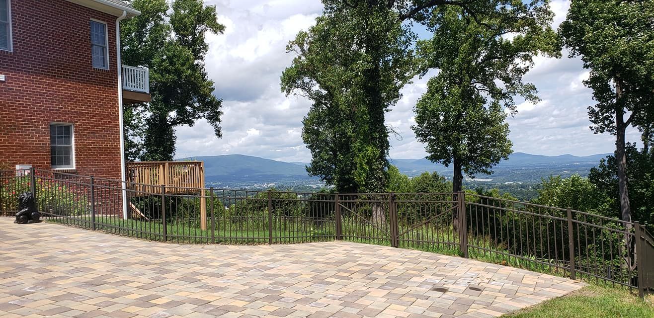 Brick house overlooking a valley, trees, and cloudy sky. Stone patio with a wrought iron fence.