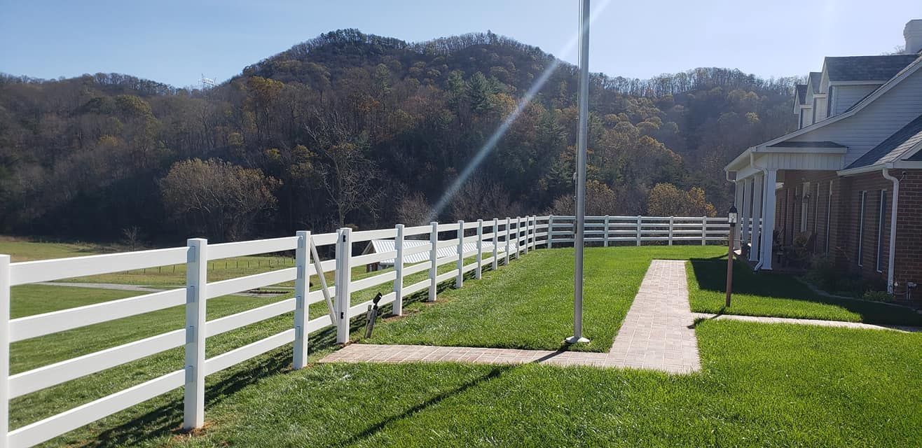 White fence with a flag pole and grass path leading to a building with a tree covered mountain in the background.