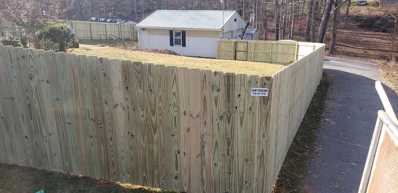 Wooden fence surrounding a small building and yard, asphalt driveway, and trees.