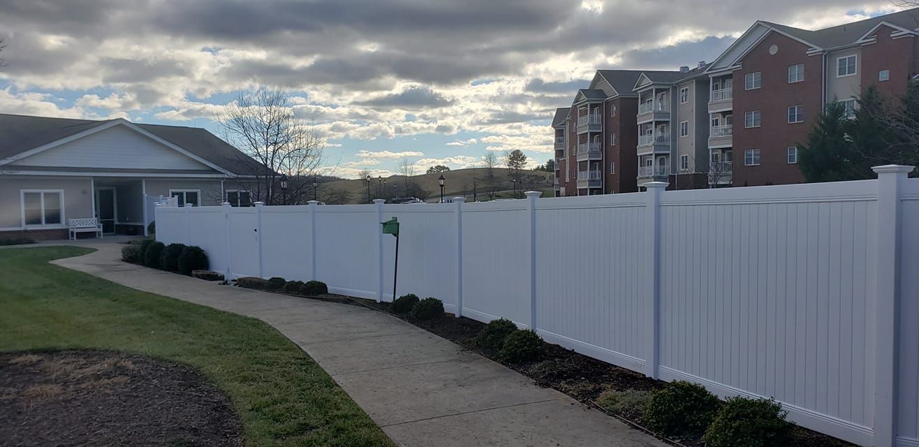 White fence bordering a paved walkway and green lawn, with a building on each side under a cloudy sky.