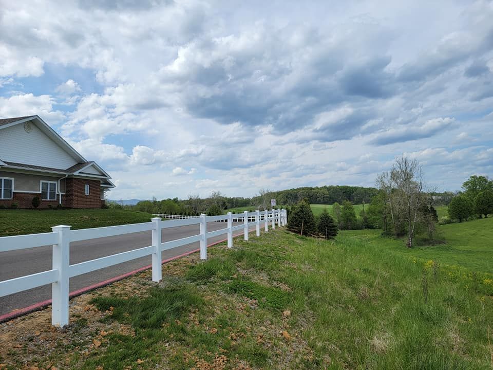 White fence alongside a road next to a green field and a brick building under a cloudy sky.