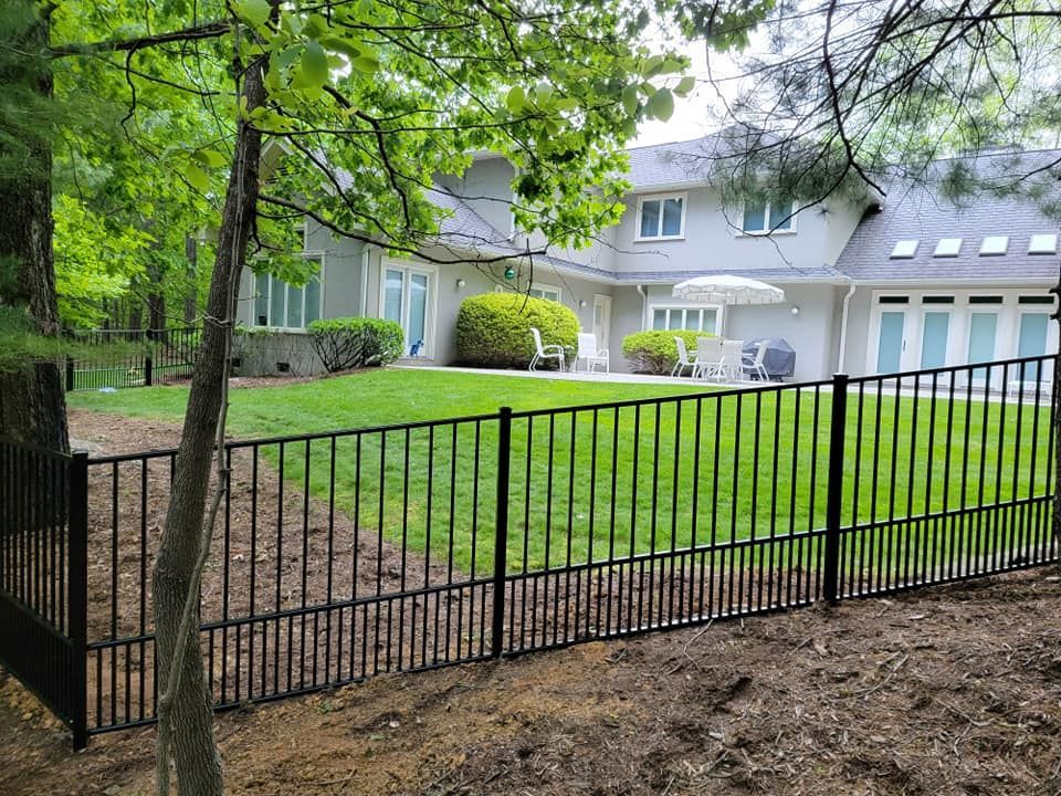 Black metal fence encloses a green lawn with a large, light-colored house in the background.