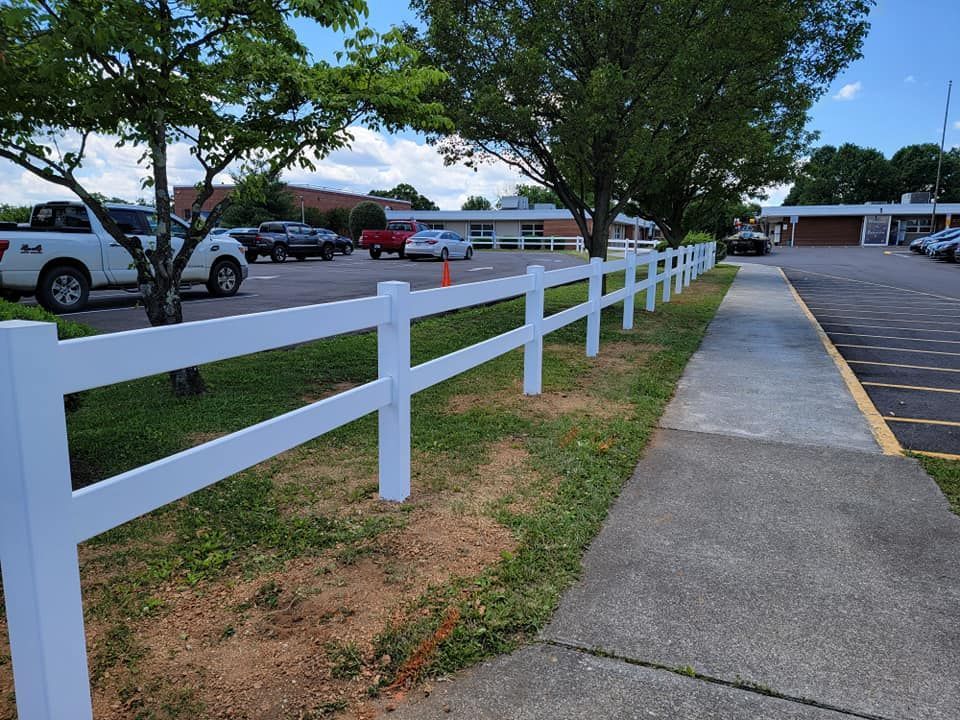 White fence bordering a sidewalk and parking lot with cars and a building in the background.