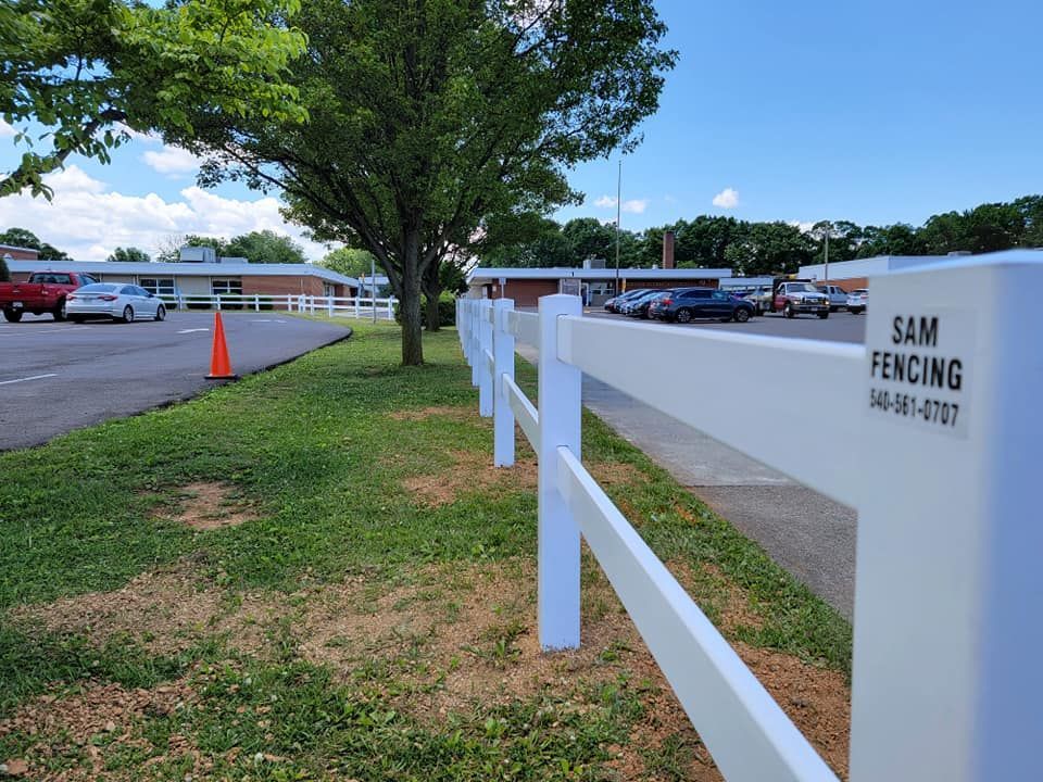 White fence along a grassy area, with a building and parked cars in the background on a sunny day.