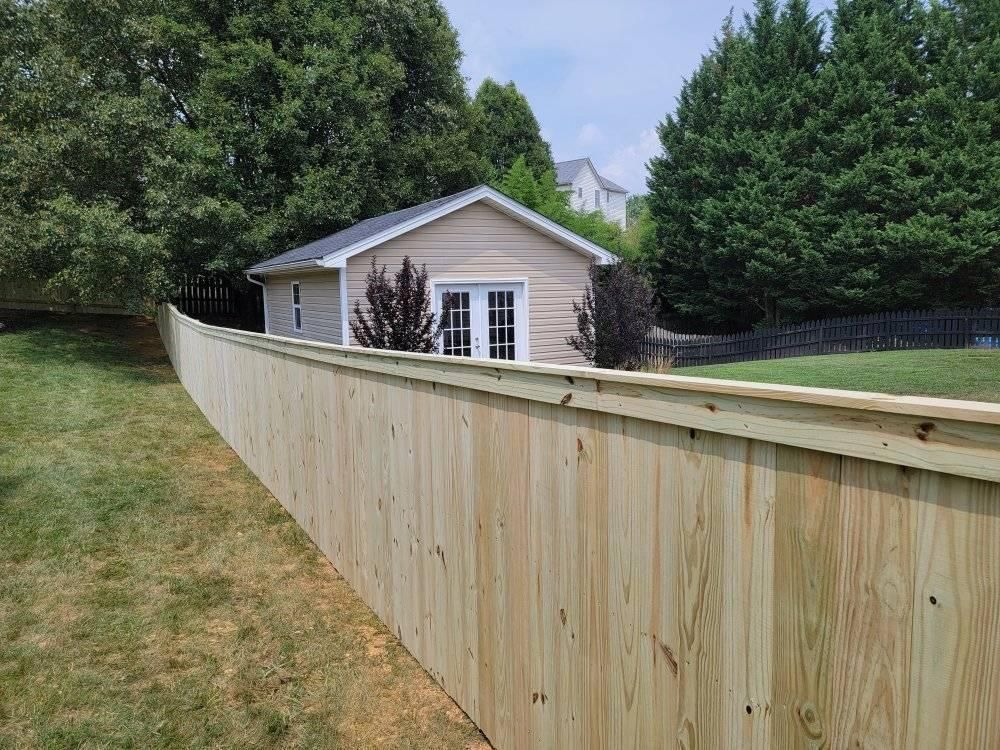 Wooden fence along a grassy slope, with a small tan building in the background surrounded by trees.