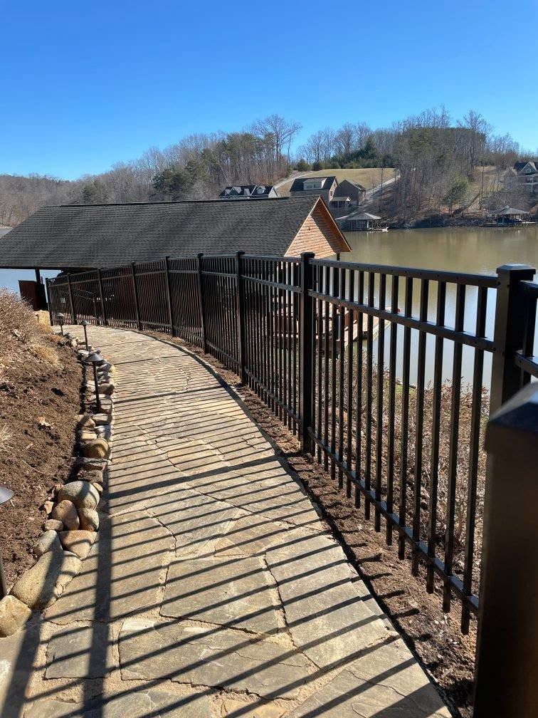 A black metal fence lines a stone walkway along the water. A boathouse is in the background.