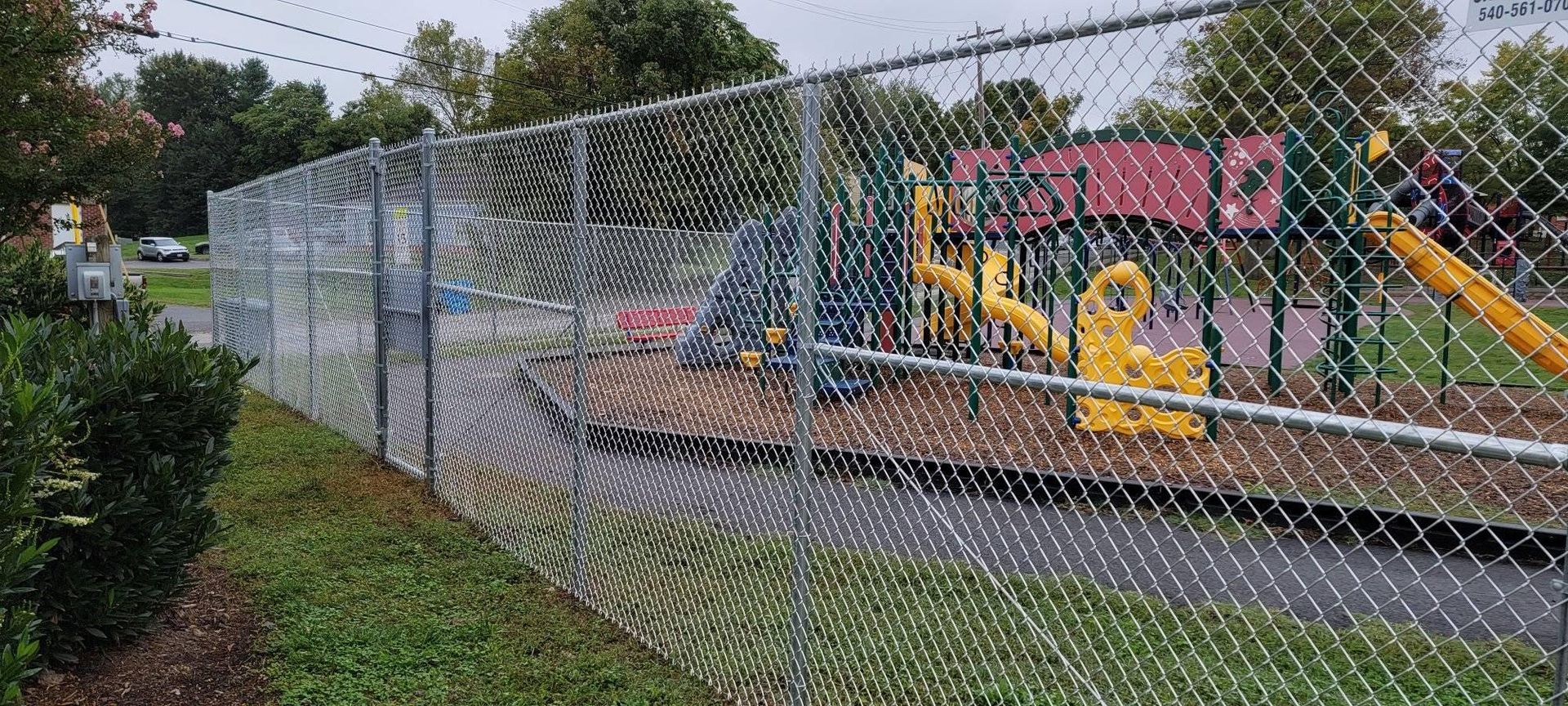 A chain link fence surrounds a playground with yellow equipment and a red structure, set on wood chips.