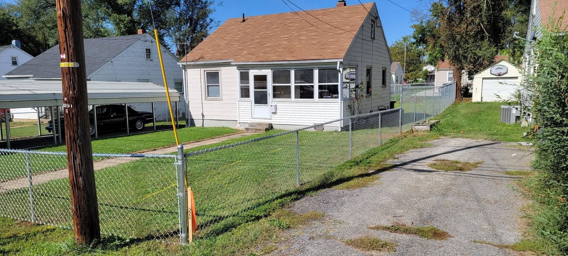 A small, light-colored house with a porch and brown roof, chain link fence, and a driveway in a residential area.