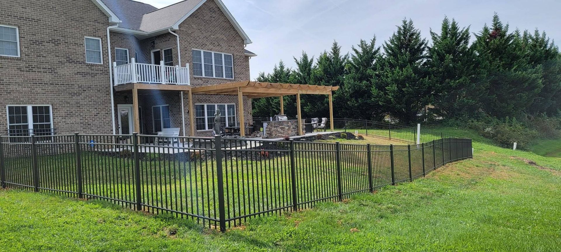A two-story brick house with a deck and pergola. A black fence surrounds the yard.
