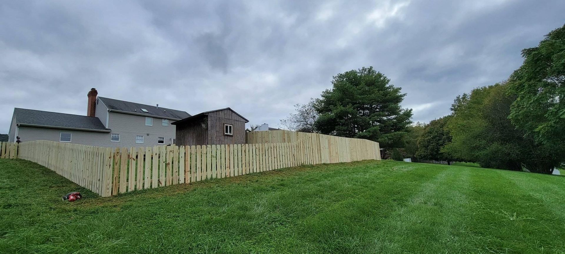 A freshly built wooden fence surrounds a grassy backyard, cloudy sky above a house.