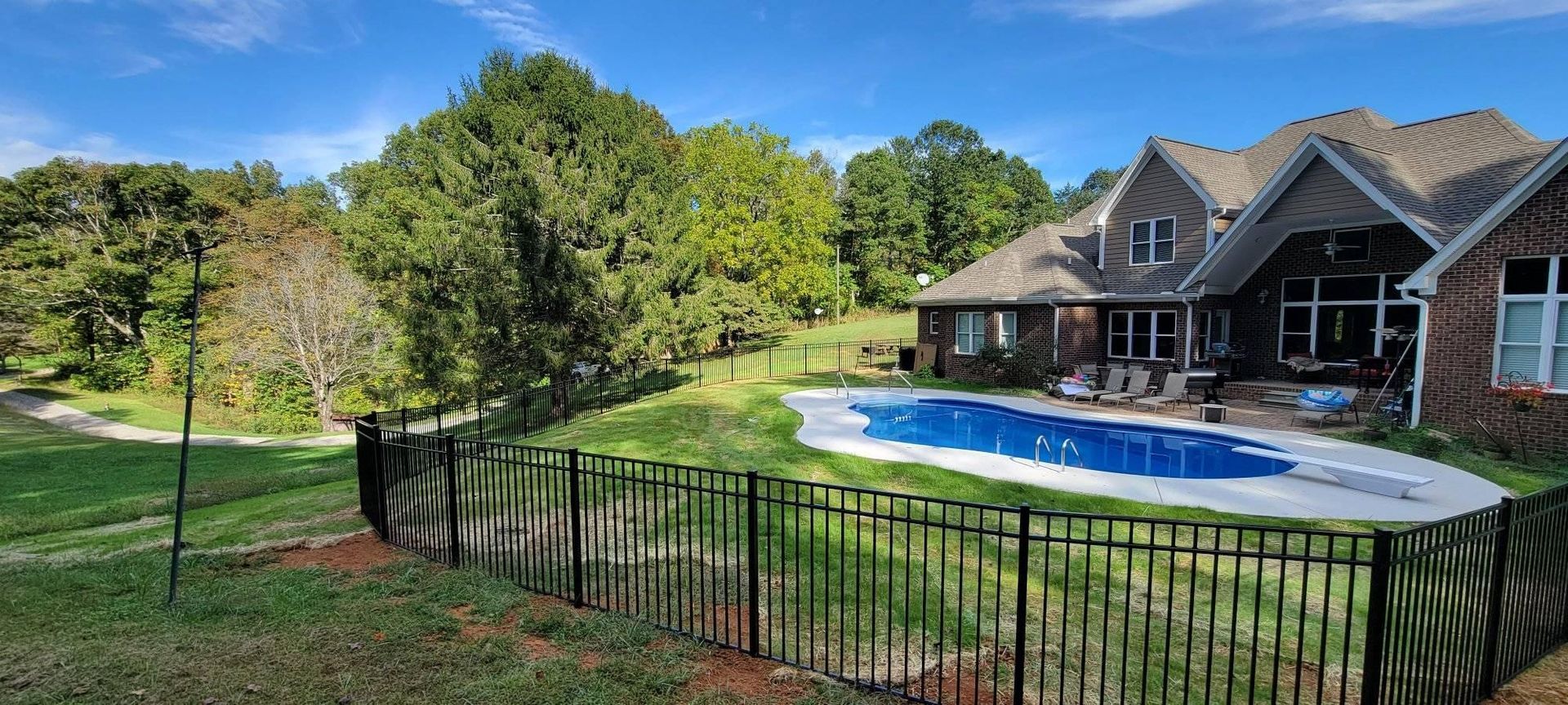 Backyard with pool, fenced area, large house, trees, and blue sky.