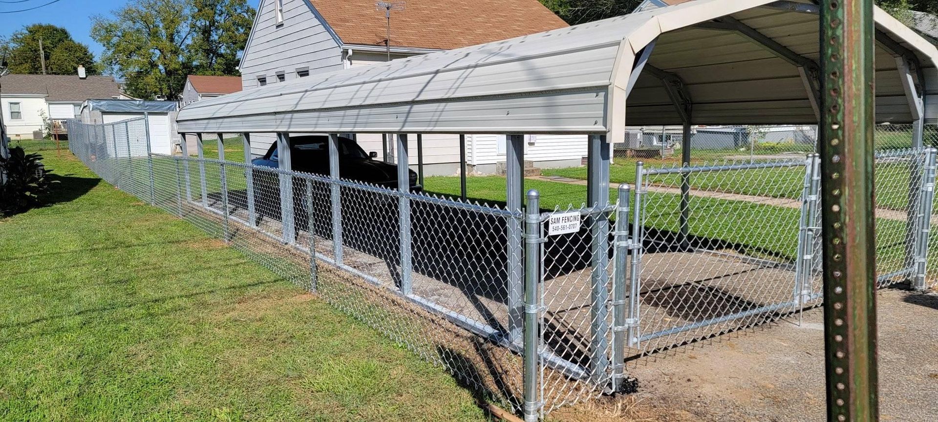 Chain-link fence surrounding a carport and grassy yard. The carport covers a car, a sign on the fence.