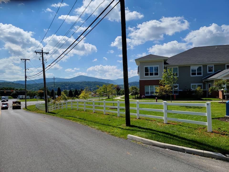 Roadside view of a building, white fence, and mountains under a blue sky with clouds. Power lines overhead.