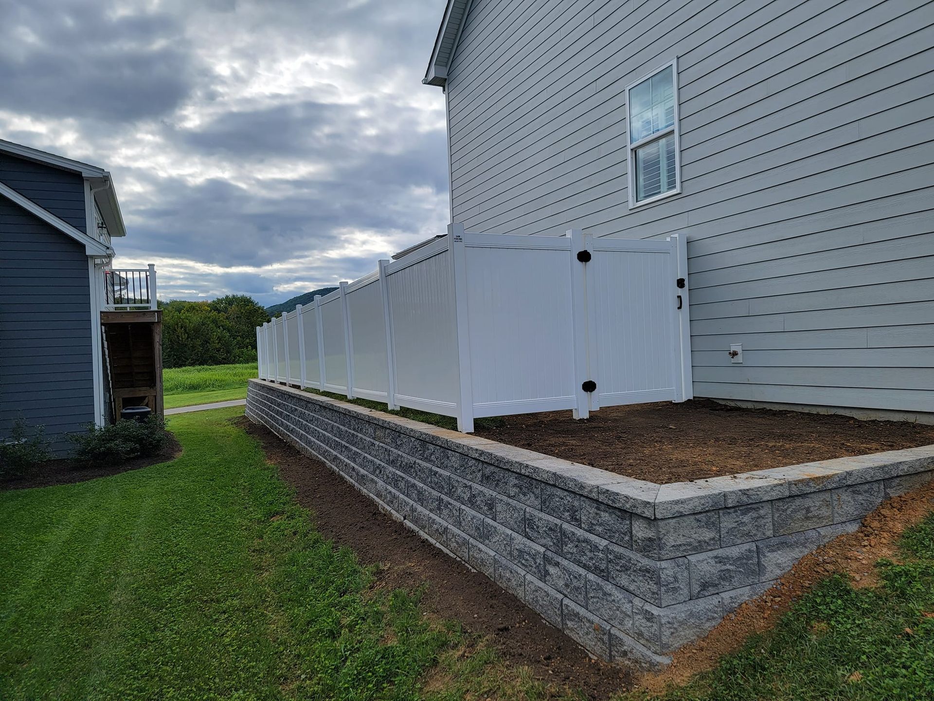 A white vinyl fence next to a building, atop a retaining wall, with a cloudy sky in the background.