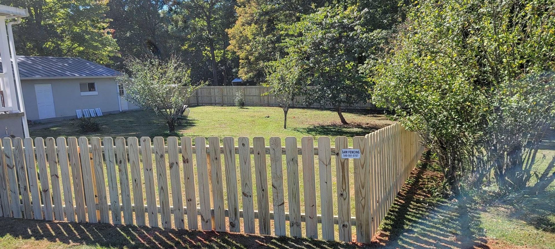 Wooden fence encloses a small yard with a building, trees, and grass.
