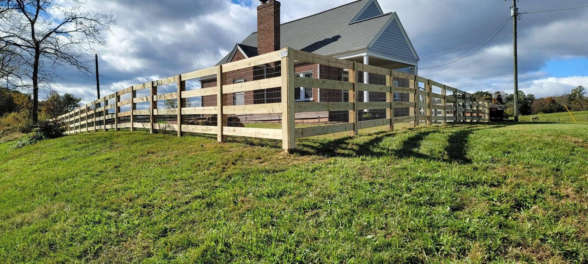 A wooden fence surrounds a house with a gray roof and brick chimney, set in a grassy field under a cloudy sky.