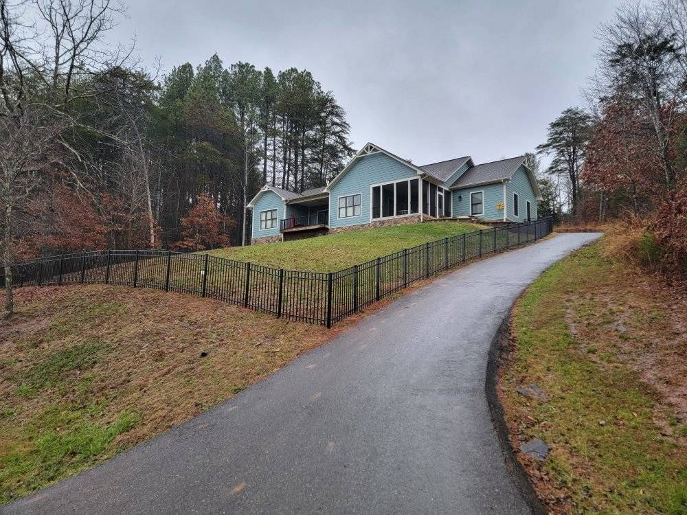 Asphalt driveway leading up to a light blue house on a hill, surrounded by a black fence and trees.
