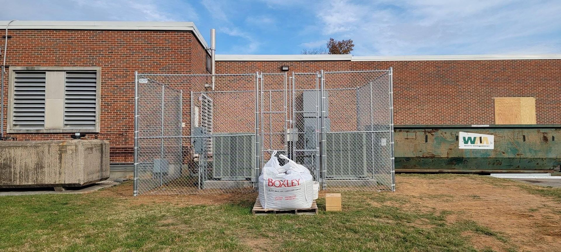 Brick building with chain link fence, HVAC units, dumpster, bag of trash, and cardboard box outside.