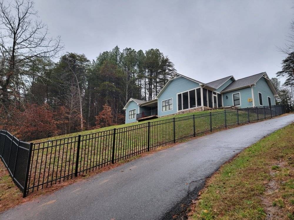 Blue house with a black fence sits on a hill beside a paved road, trees in the background.