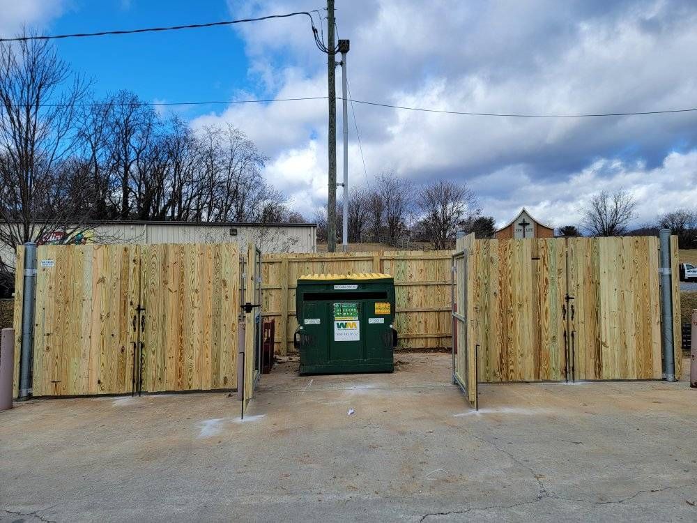 Wooden fence encloses a green dumpster on a concrete pad. Gates are open. Cloudy sky.