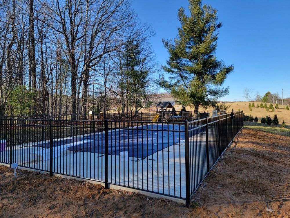 Black fence surrounds a rectangular pool covered with a blue tarp, set in a yard with trees and a clear blue sky.