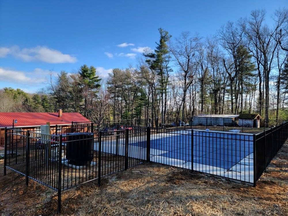A black fenced-in pool with a blue cover, with surrounding trees and a red-roofed building.