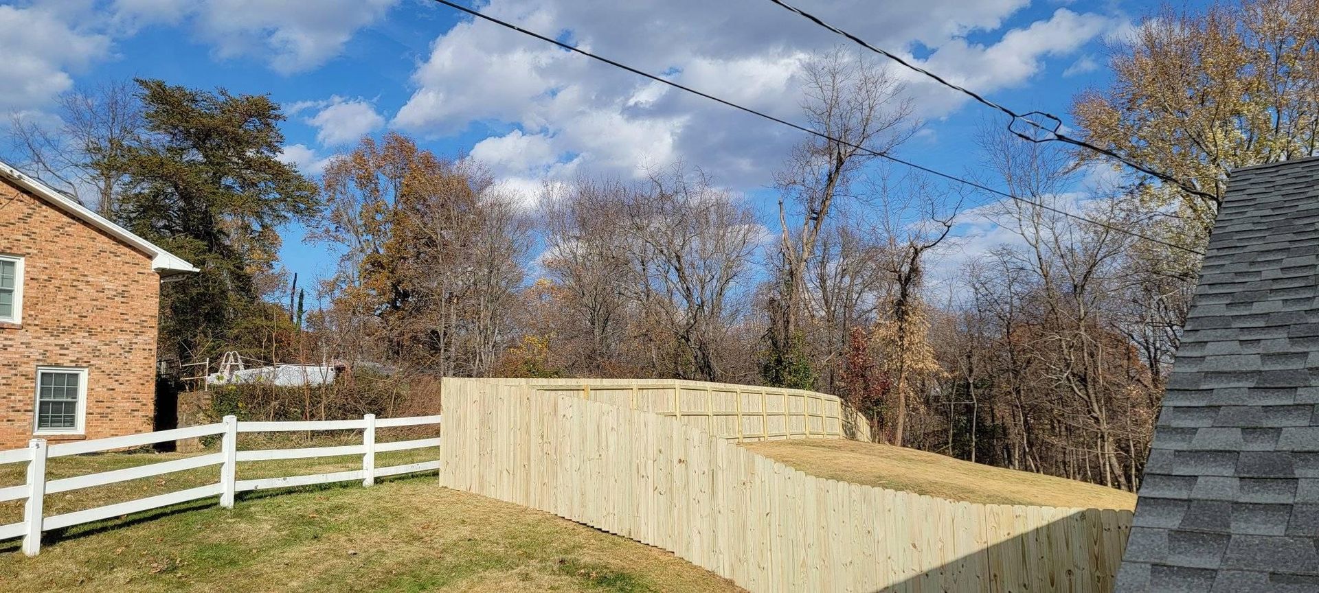 A view of a building, a white fence, a wooden fence, and trees under a blue sky.