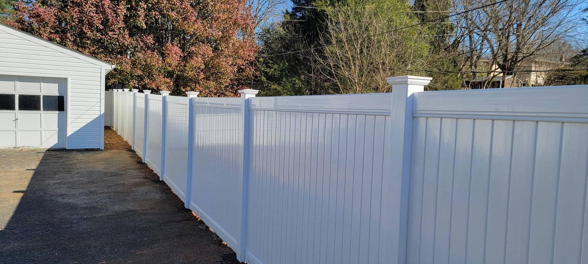 White fence along a gravel driveway next to a white garage. Trees in background.