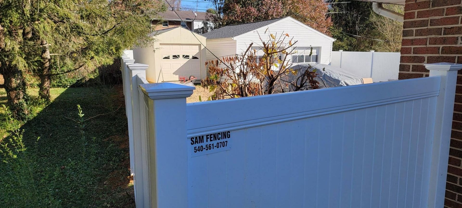 White fence with some brown stains in front of a house, yard with trees, and a beige garage.