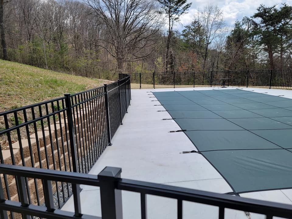 Black fence surrounds a pool with a green cover, set against a backdrop of trees and a hillside.