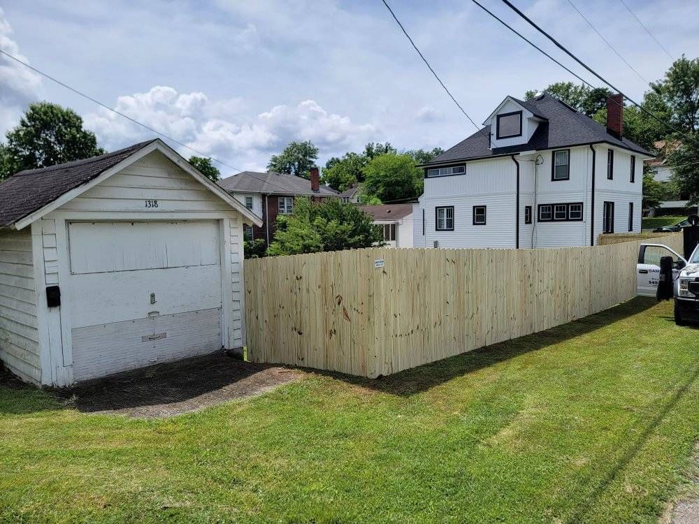 Wooden fence in a front yard, with a white garage and house visible. Green grass and blue sky.