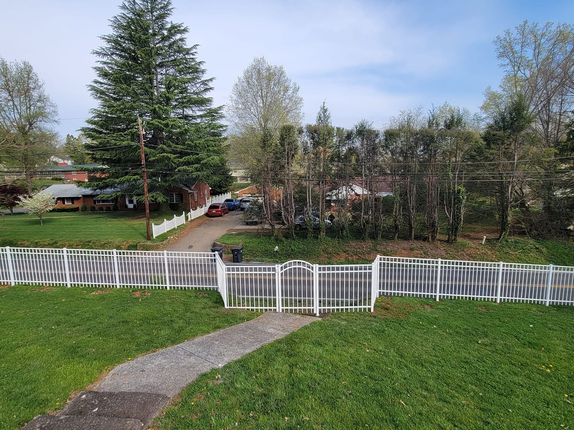 White picket fence with gate, gravel path, green grass, trees, and houses in the background.