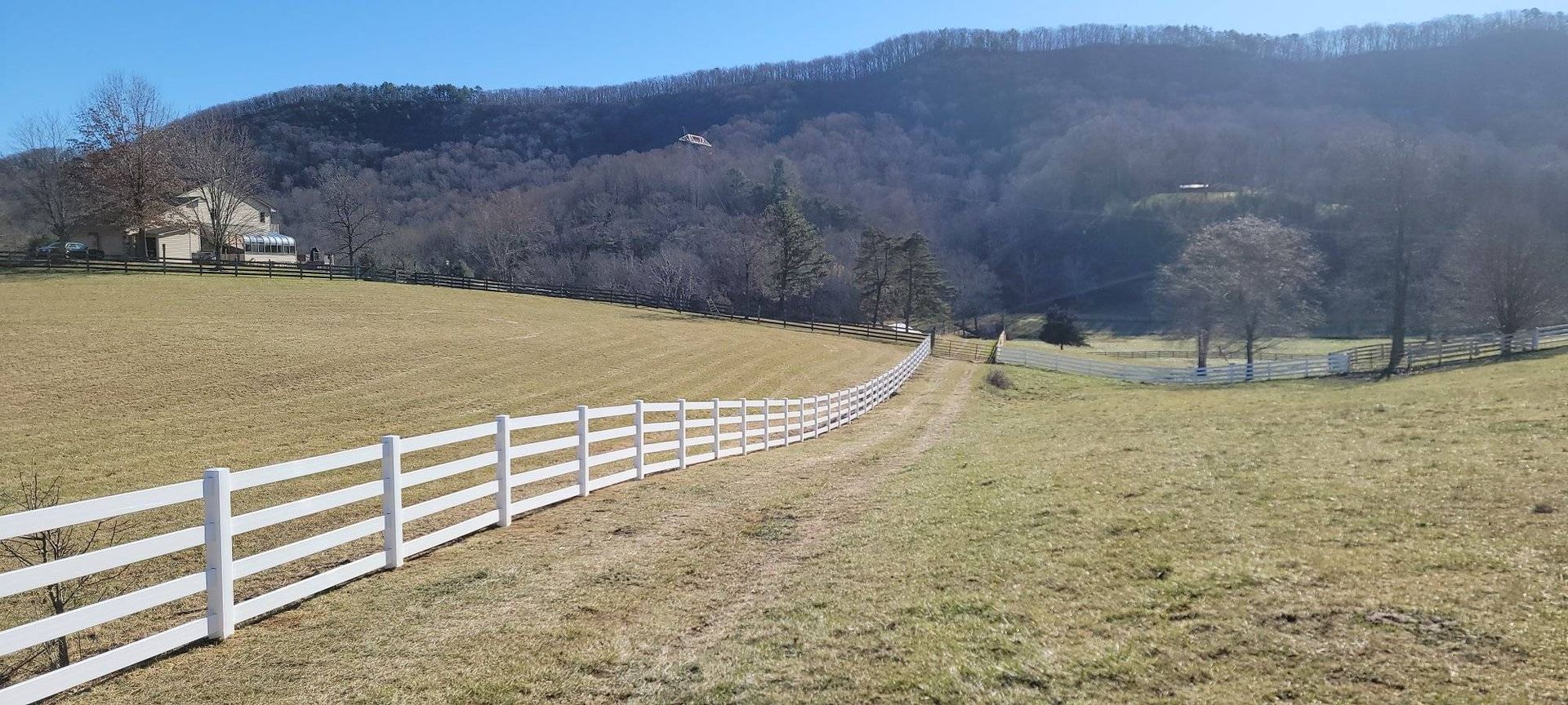 White fence in a grassy field, leading towards a tree-covered mountain under a blue sky.
