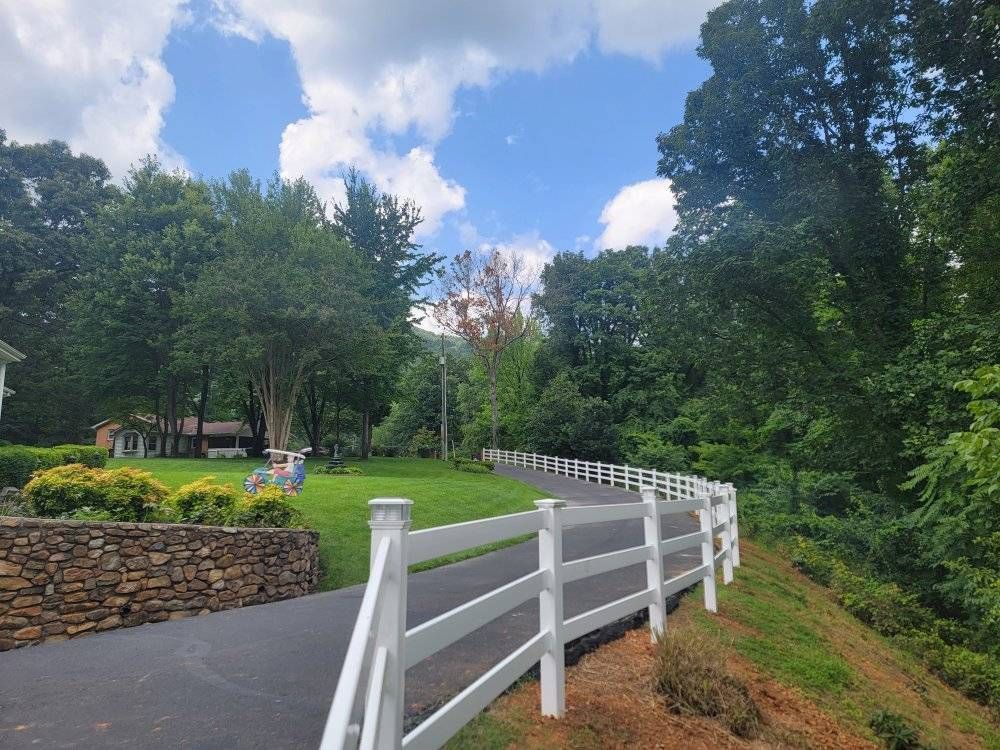 A paved driveway with a white fence alongside green grass and trees under a blue sky.