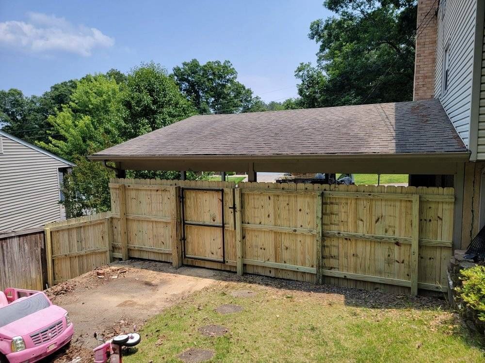 Wooden fence enclosing a small backyard area with a gate, adjacent to a house with a carport roof.
