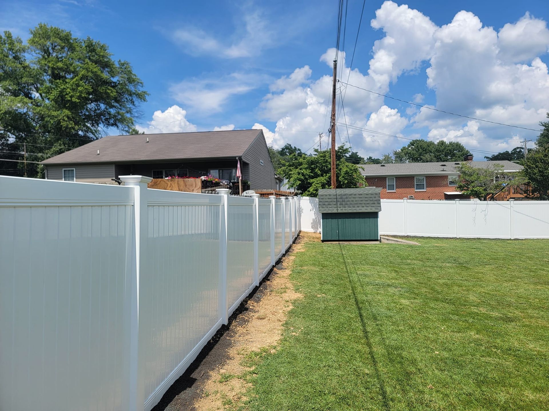 White vinyl fence in a grassy backyard, adjacent to houses under a partly cloudy blue sky.