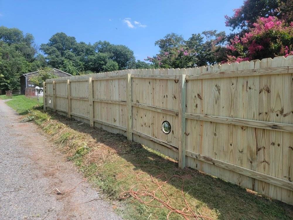 Wooden fence along a gravel path and grassy area, under a blue sky, with trees in the background.