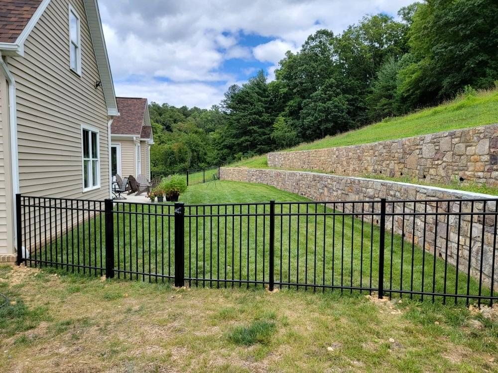 Black metal fence encloses a grassy yard next to a beige house, with terraced stone retaining walls and trees in the background.