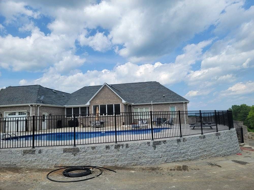 House with swimming pool behind a black fence and retaining wall under a cloudy sky.