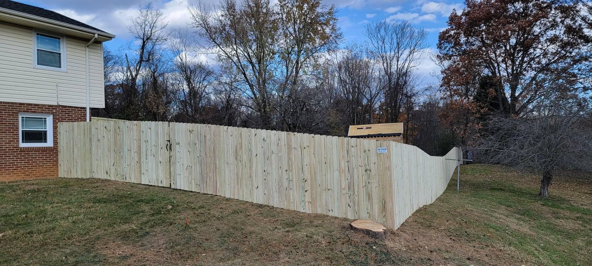 A wooden fence curves along a grassy yard next to a house under a partly cloudy sky.