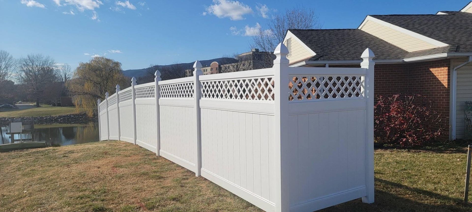 White fence along a grassy area next to a body of water and building under a blue sky.