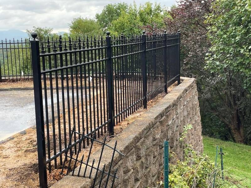 Black metal fence atop a stone wall, bordering a grassy area and road, with trees in the background.