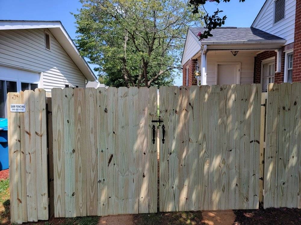 Wooden fence gate with black hardware in front of houses on a sunny day.